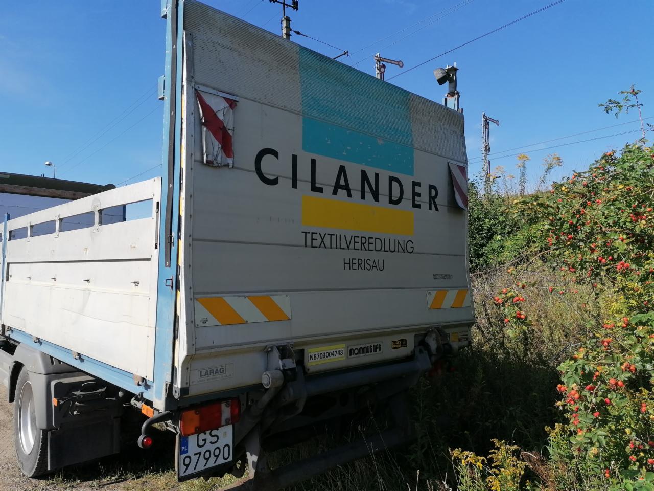 Ciężarówka z otwartą skrzynią ładunkową, widoczne logo firmy Cilander Textilveredlung Herisau na boku, zaparkowana na poboczu z bujną roślinnością.