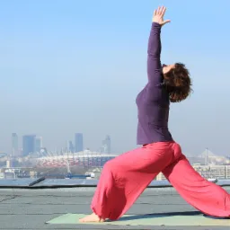Kobieta w pozycji wojownika na macie do jogi na dachu z widokiem na panoramę Warszawy w tle.
