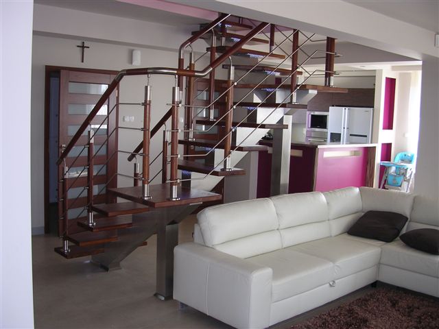 Nowoczesne schody drewniane z metalowymi balustradami i podświetleniem LED, widoczne z perspektywy salonu z białym narożnikiem i brązowym dywanem.