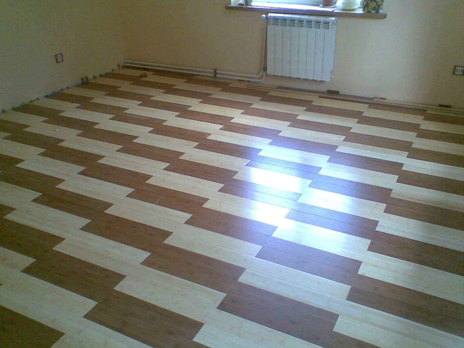 Ukończone układanie dwukolorowych paneli podłogowych w jodełkę, widoczne listwy przypodłogowe w trakcie montażu, grzejnik na ścianie.