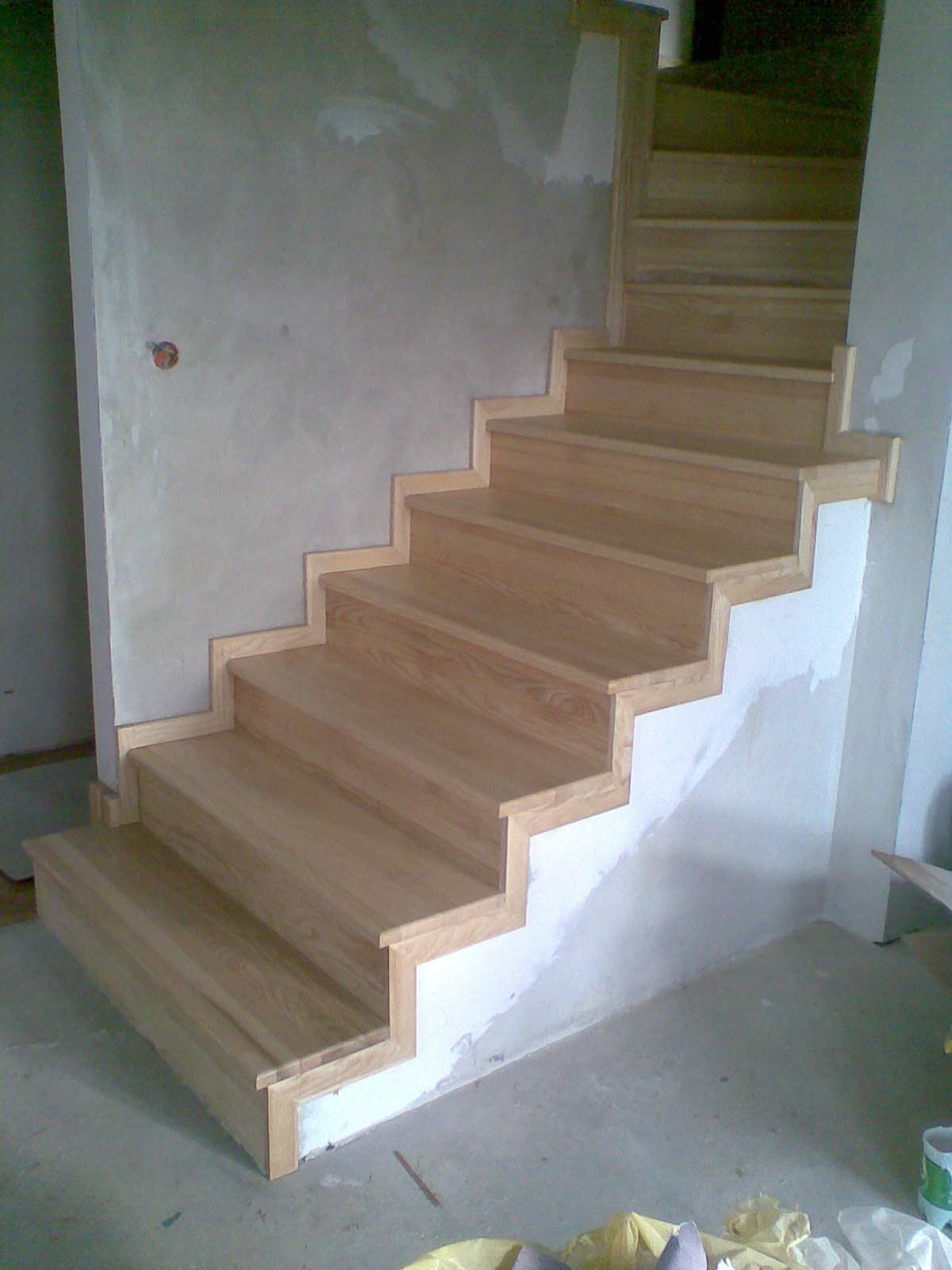Drewniane schody z jasnego drewna w trakcie montażu, widoczne niedoskonałości na ścianie i podłodze, surowy stan wykończenia wnętrza.