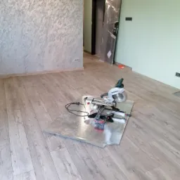 Pokój w trakcie remontu z nową podłogą z paneli, na której stoi ukośnica na blasze ochronnej, ściana w tle z dekoracyjną fakturą.