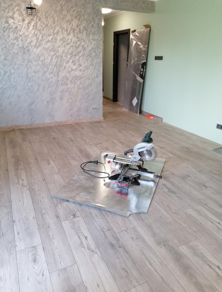 Pokój w trakcie remontu z nową podłogą z paneli, na której stoi ukośnica na blasze ochronnej, ściana w tle z dekoracyjną fakturą.