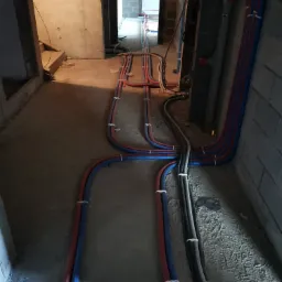 Wnętrze w trakcie remontu z widocznymi rurami instalacji wodnej i grzewczej, ułożonymi na podłodze i przymocowanymi do ściany, prowadzącymi przez surowy stan pomieszczeń.