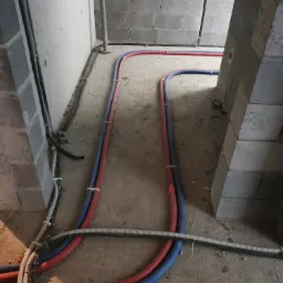 Instalacja podłogowa ogrzewania rurami PEX w kolorze czerwonym i niebieskim, przymocowanymi do podłoża w surowym stanie, widoczne szare bloczki betonowe ścian i słupów.