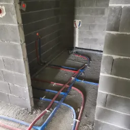 Surowe wnętrze łazienki w trakcie prac instalacyjnych; widoczne szare ściany z bloczków betonowych, czerwone i niebieskie rury instalacji wodnej ułożone na podłodze, oraz wystające przyłącza...