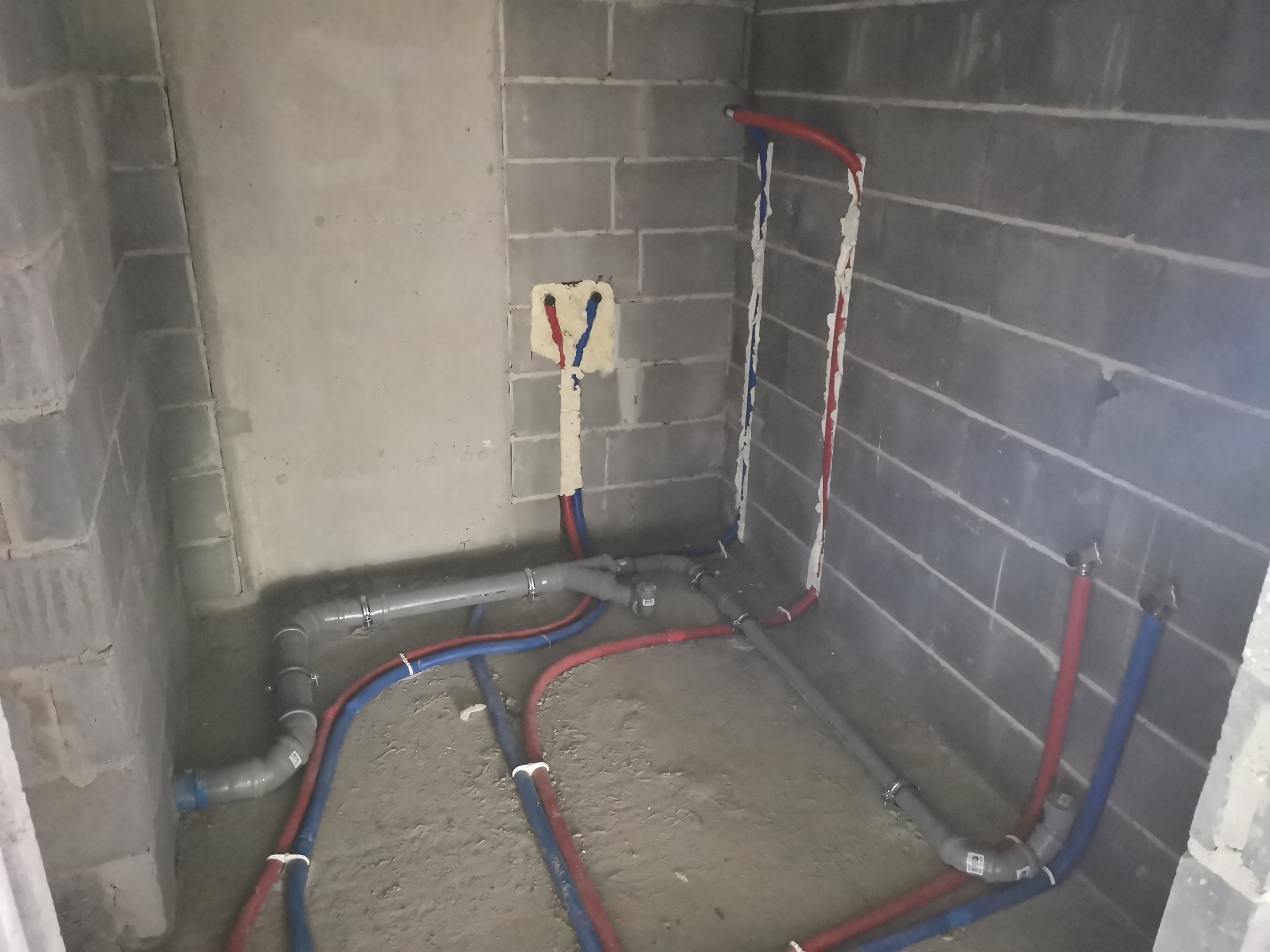 Surowy stan łazienki z widocznymi rurami instalacji wodnej i kanalizacyjnej w kolorach niebieskim i czerwonym, częściowo zabezpieczonymi pianką montażową na ścianie z szarego betonu komórkowego.