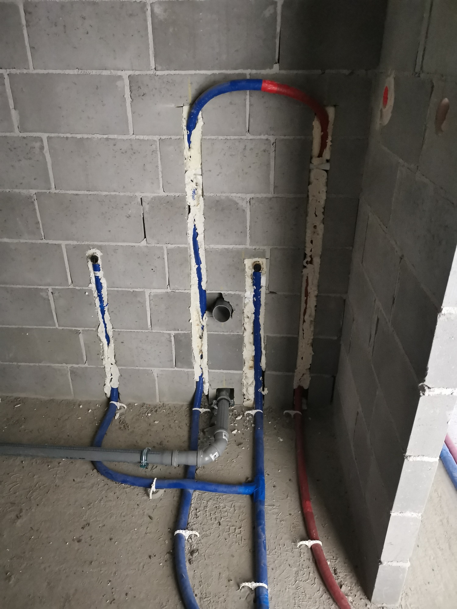 Instalacja hydrauliczna i grzewcza w surowym stanie: niebieskie i czerwone rury prowadzone w bruzdach w ścianie z bloczków, zabezpieczone pianką montażową, widoczne elementy kanalizacji.