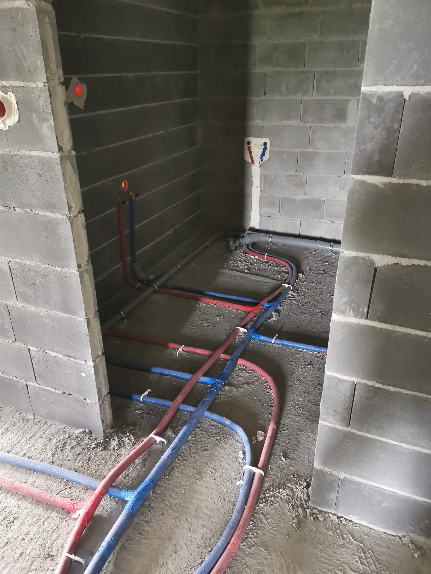 Surowe wnętrze łazienki w trakcie prac instalacyjnych; widoczne szare ściany z bloczków betonowych, czerwone i niebieskie rury instalacji wodnej ułożone na podłodze, oraz wystające przyłącza...