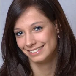 Portret uśmiechniętej brunetki z niebieskimi oczami z Krakowa, z delikatnym makijażem i błyszczykiem na ustach, na neutralnym tle.