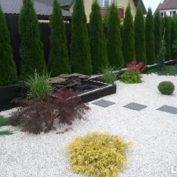 Nowoczesny ogr&oacute;d z białym żwirem, rzędem tuj wzdłuż ogrodzenia, małym oczkiem wodnym z kaskadą z kamienia łupkowego oraz ścieżką z płyt chodnikowych.