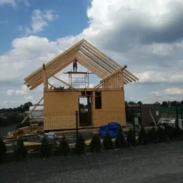 Wznoszenie domu szkieletowego: konstrukcja z drewnianych belek i płyt OSB, pracownik na rusztowaniu na tle błękitnego nieba z chmurami, widok z poziomu gruntu.