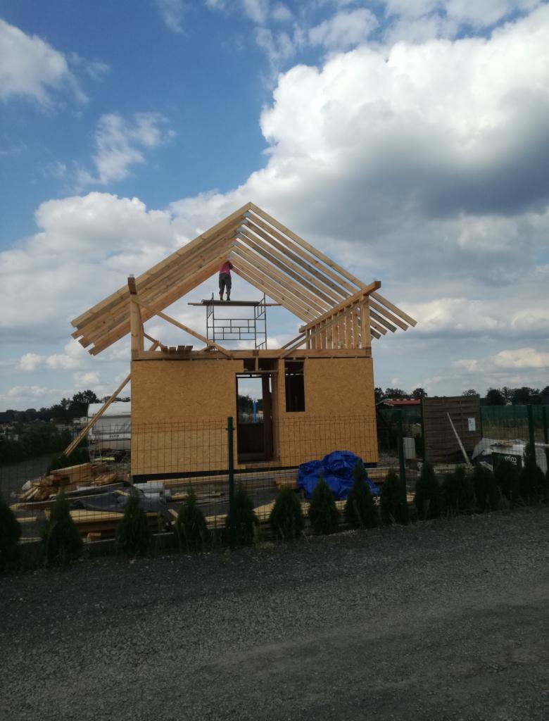 Wznoszenie domu szkieletowego: konstrukcja z drewnianych belek i płyt OSB, pracownik na rusztowaniu na tle błękitnego nieba z chmurami, widok z poziomu gruntu.