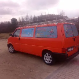 Pomarańczowy samochód dostawczy z reklamą usług hydraulicznych 'HydroKan' na bocznych szybach, zaparkowany na żwirowej powierzchni z widocznym bagażnikiem dachowym i numerem telefonu.