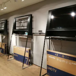 Ekspozycja trzech telewizorów Samsung UHD TV 55 cali w czarnych, minimalistycznych ramach z półkami w sklepie, opakowania pod spodem, oświetlenie sufitowe.