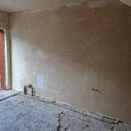 Pokój w trakcie remontu: ściany pokryte świeżym tynkiem w odcieniach beżu, podłoga z pozostałościami kleju po płytkach, widoczne rury instalacyjne i skrzynka rewizyjna.