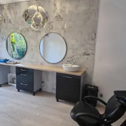 Nowoczesna łazienka z marmurową ścianą, okrągłymi lustrami i designerskimi lampami wiszącymi; widoczne czarne szafki pod umywalką i fotel fryzjerski sugerują adaptację przestrzeni na salon.