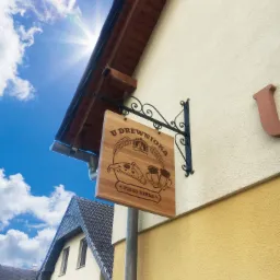 Drewniany szyld restauracji 'U Drewnioka' z grafiką pizzy, piwa i zboża, zawieszony na ozdobnym, kutym wsporniku na elewacji budynku, widoczny z dołu na tle błękitnego nieba.