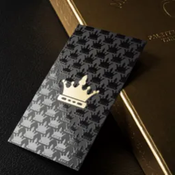 Elegancka wizytówka z tłoczonym wzorem koron na szarym tle, z pojedynczą, złotą koroną na pierwszym planie, leżąca obok złotego pudełka z wytłoczonym napisem na czarnym, skórzanym tle.