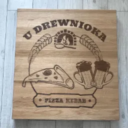Drewniana tablica z wypalonym laserowo logo pizzerii 'U DREWNIOKA', przedstawiająca piec, kłosy zboża, kufle piwa i pizzę, umieszczona na podłodze z paneli.