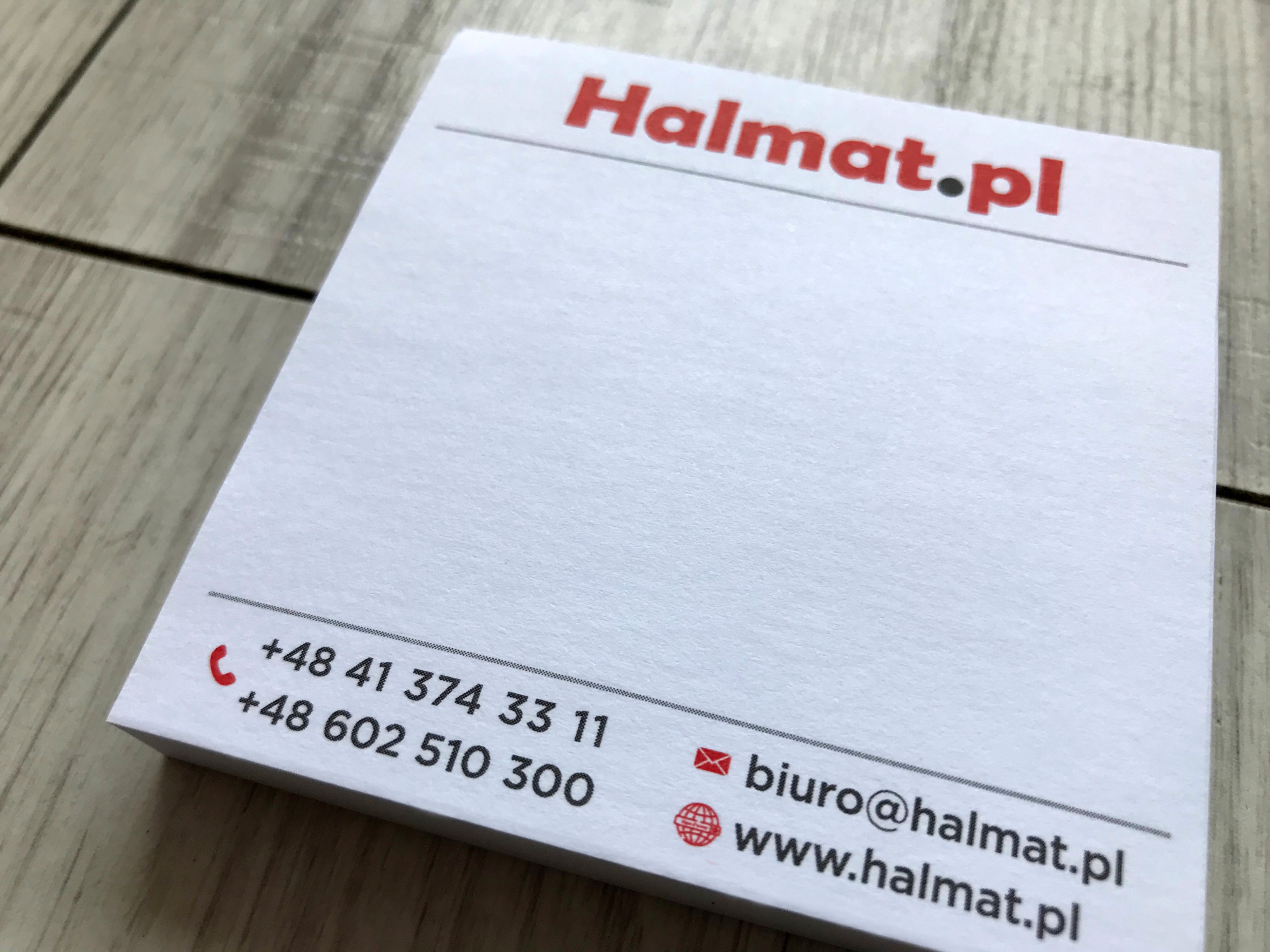 Blok kartek biurowych z nadrukowanym logo Halmat.pl, numerami telefonów i adresem e-mail, leżący na drewnianym stole.