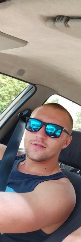 Mężczyzna w okularach przeciwsłonecznych siedzi w samochodzie z zapiętym pasem bezpieczeństwa. Widoczny fragment deski rozdzielczej i otoczenie za oknem.