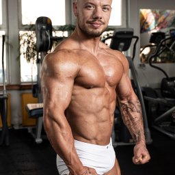 Bartłomiej Chojowski TRENER PERSONALNY - Umięśniony mężczyzna z tatuażami na ramionach i dłoniach, pozujący w białej bieliźnie na tle siłowni z maszynami do ćwiczeń.