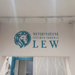 Niebieski logotyp 'Weterynaryjne Centrum Zdrowia LEW' z symbolem lwa na białej ścianie, oświetlony reflektorami sufitowymi i monitorowany przez kamerę.