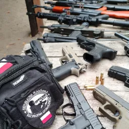 Różne modele broni palnej, karabinów i pistoletów ułożone na drewnianym stole, obok torby z naszywką z motywem strzelca i flagą Polski.