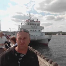 Mężczyzna na molo z białym statkiem pasażerskim 'Rubin' w tle, pochmurne niebo, widok na plażę i budynki w oddali.