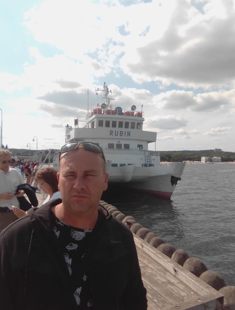 Mężczyzna na molo z białym statkiem pasażerskim 'Rubin' w tle, pochmurne niebo, widok na plażę i budynki w oddali.