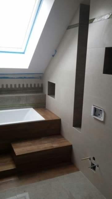 Wykończona łazienka na poddaszu z obudowaną wanną drewnopodobnymi płytkami, szara glazura ścienna z wnękami i oknem dachowym.