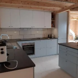 Jasna kuchnia z szarymi szafkami i czarnym blatem, widoczne drewniane elementy konstrukcyjne domu, sprzęt pomiarowy na pierwszym planie.