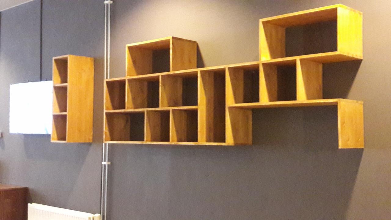 Drewniane, modułowe półki ścienne o nieregularnym kształcie, zamontowane na szarej ścianie, pojedyncza kolumna z półkami po lewej stronie.