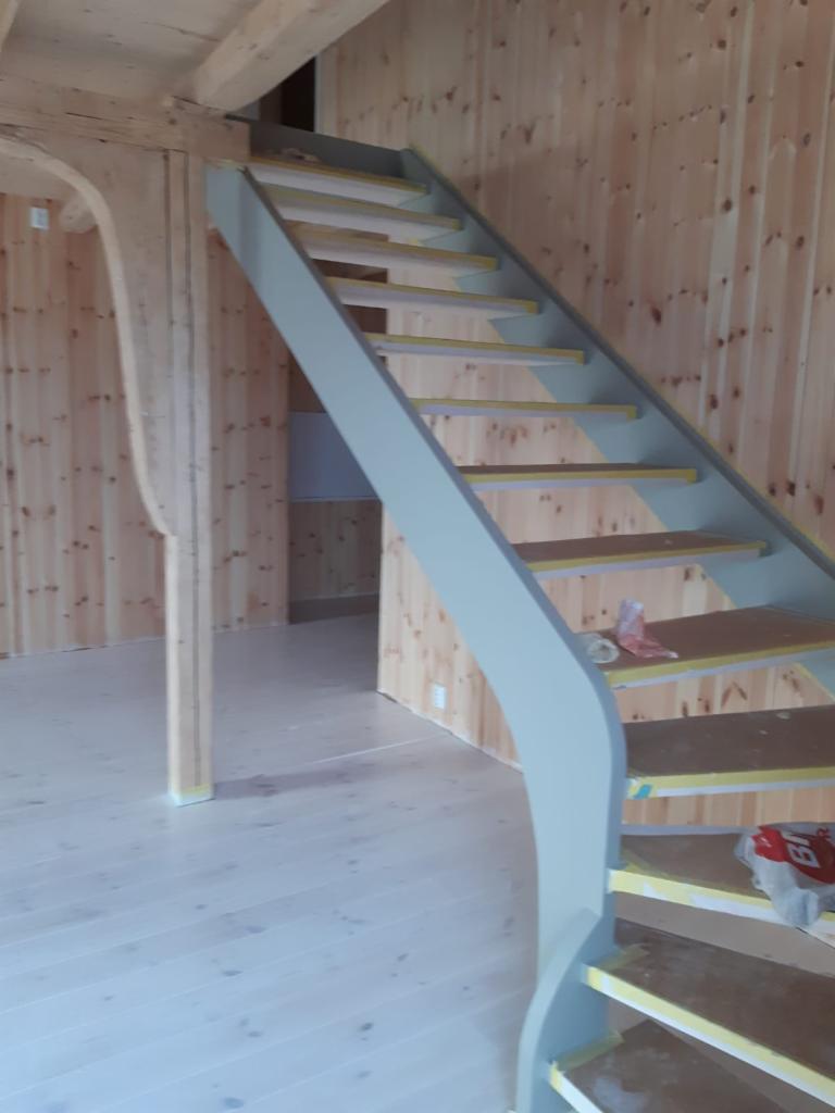 Drewniane schody z szarymi elementami w surowym wnętrzu domu w trakcie wykańczania, widoczne zabezpieczenia taśmą malarską i resztki materiałów budowlanych.