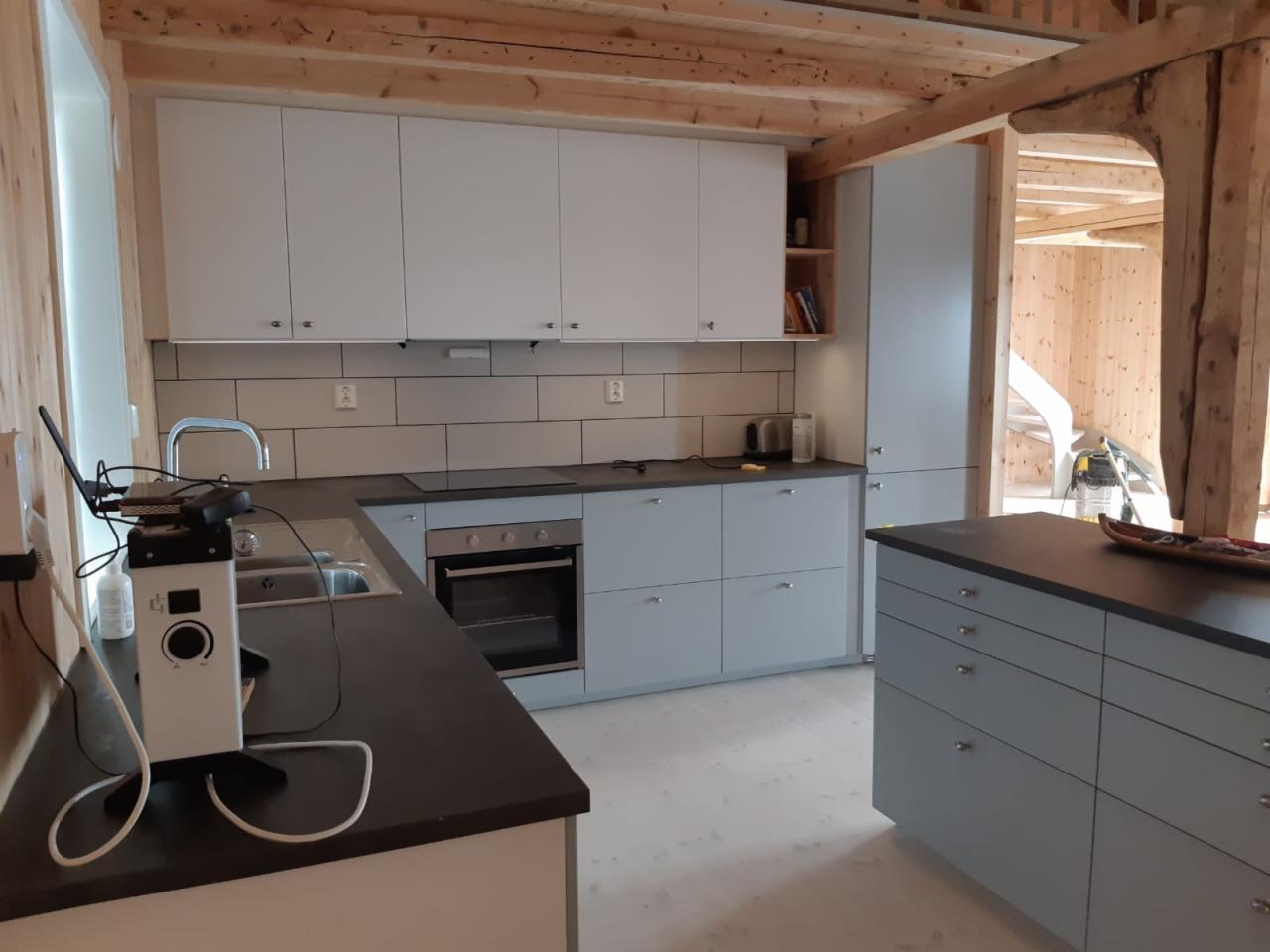 Jasna kuchnia z szarymi szafkami i czarnym blatem, widoczne drewniane elementy konstrukcyjne domu, sprzęt pomiarowy na pierwszym planie.