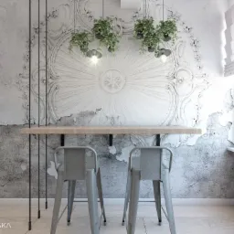 Biała ściana z dekoracyjnym ornamentem i efektem spękań, drewniany blat barowy z dwoma metalowymi hokerami, wiszące rośliny i lampy, widok na miasto z okna.
