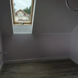 Poddasze po remoncie: pomalowane ściany w jasnym odcieniu fioletu, drewnopodobna podłoga, okno dachowe z widokiem na dom i zieleń, widoczne gniazdko elektryczne z wystającymi przewodami.