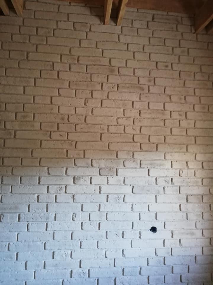 Ściana z białej cegły dekoracyjnej z widocznym otworem na gniazdko elektryczne i drewnianym stropem w tle.