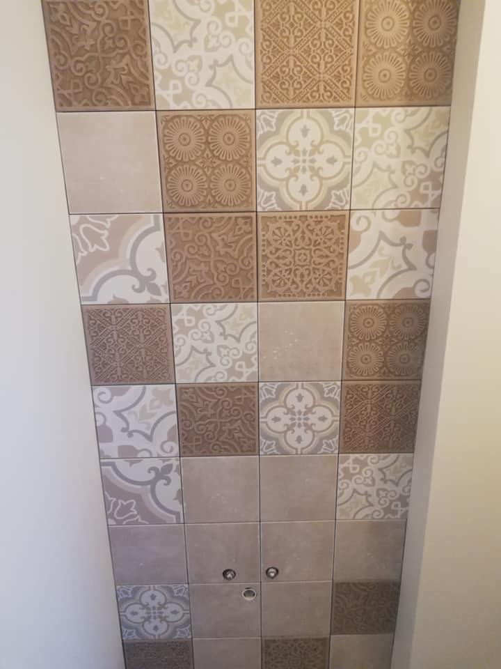 Fragment ściany wykończonej kwadratowymi płytkami ceramicznymi w odcieniach beżu i brązu z różnorodnymi wzorami, widoczne dwa okrągłe elementy instalacji.