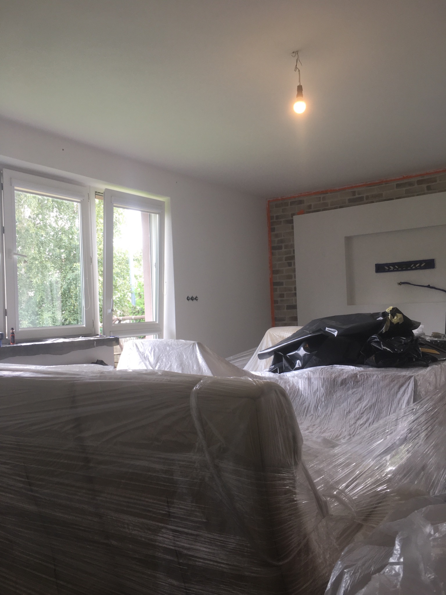 Przygotowanie do prac wykończeniowych: meble zabezpieczone folią malarską w jasnym pomieszczeniu z oknem i surową cegłą na ścianie, widoczna żarówka na suficie.