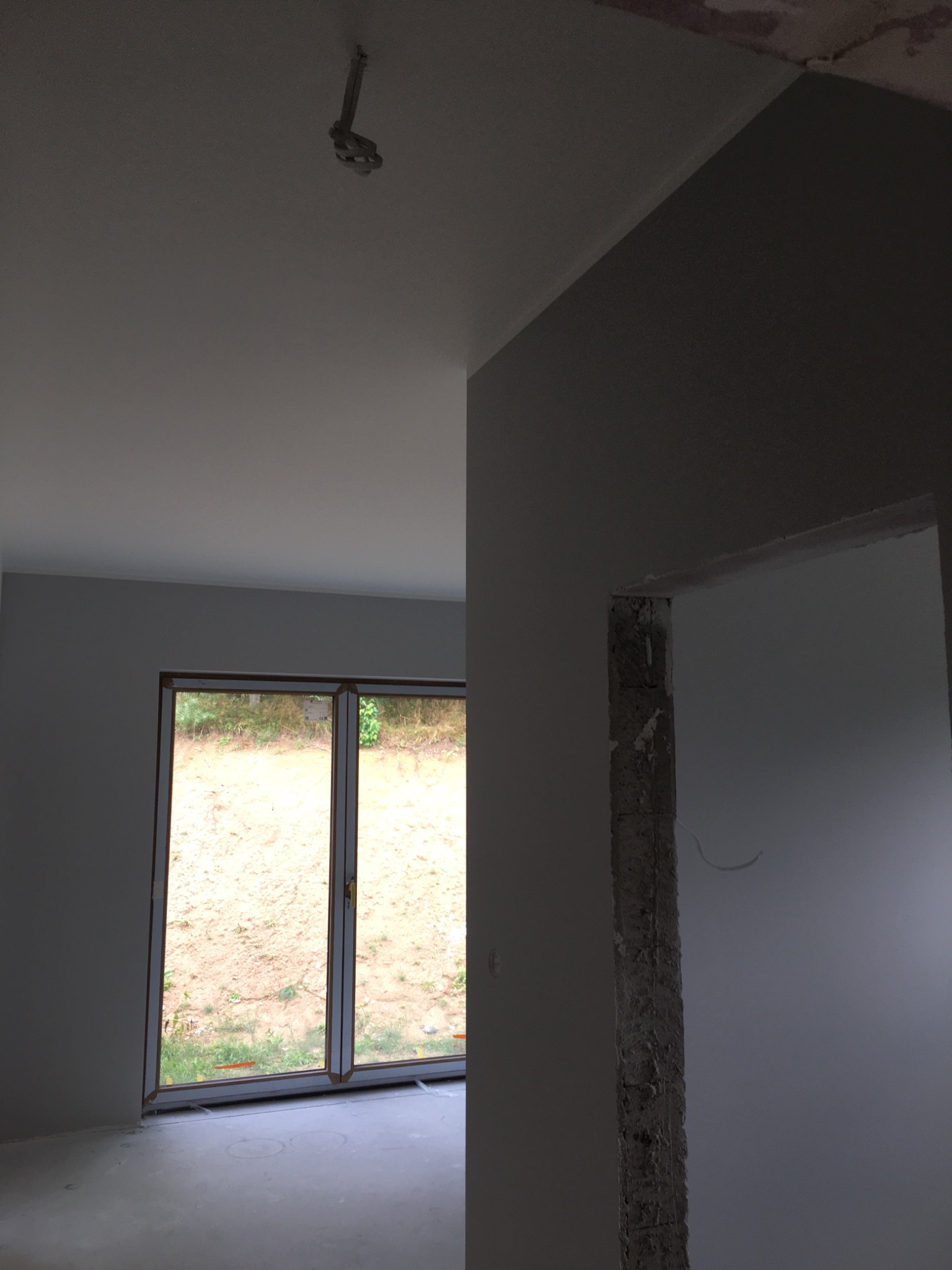 Wnętrze w trakcie malowania na szaro, widoczne okno balkonowe i surowa ościeżnica drzwiowa, z wystającym kablem elektrycznym pod sufitem.