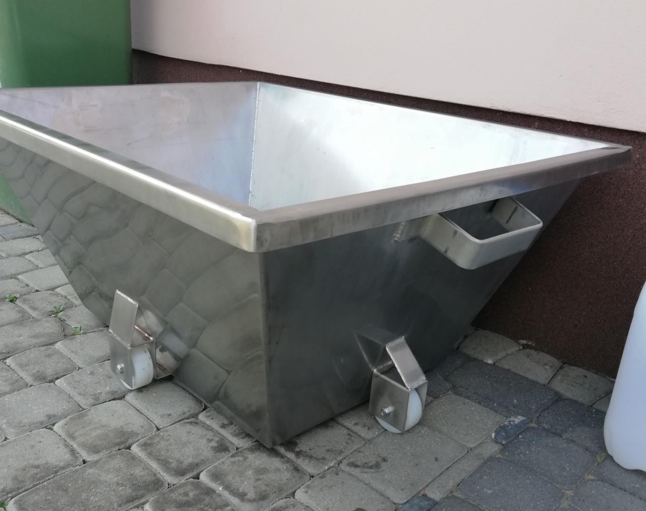 Metalowy kontener na kółkach, spawany z blachy nierdzewnej, widoczne spawy i uchwyty, stoi na kostce brukowej.