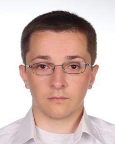 Portret młodego mężczyzny w okularach, na białym tle, w jasnej koszuli. Ujęcie od przodu, neutralny wyraz twarzy.