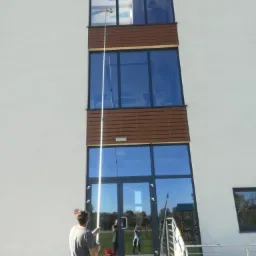 Pracownik myje okna na wysokości w budynku biurowym za pomocą teleskopowego kija z końcówką do mycia, widoczne odbicie otoczenia w oknach, czerwone wiadro na schodach.