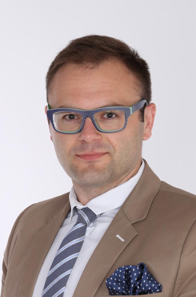 Portret mężczyzny w okularach w paski, brązowej marynarce i krawacie w paski na białym tle. Widoczna poszetka w niebieskie kropki w kieszonce marynarki.