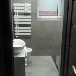 Nowoczesna, szara łazienka z grzejnikiem drabinkowym, umywalką nablatową i widokiem z okna na budynek z ceglanym dachem, widziana zza uchylonych drzwi.