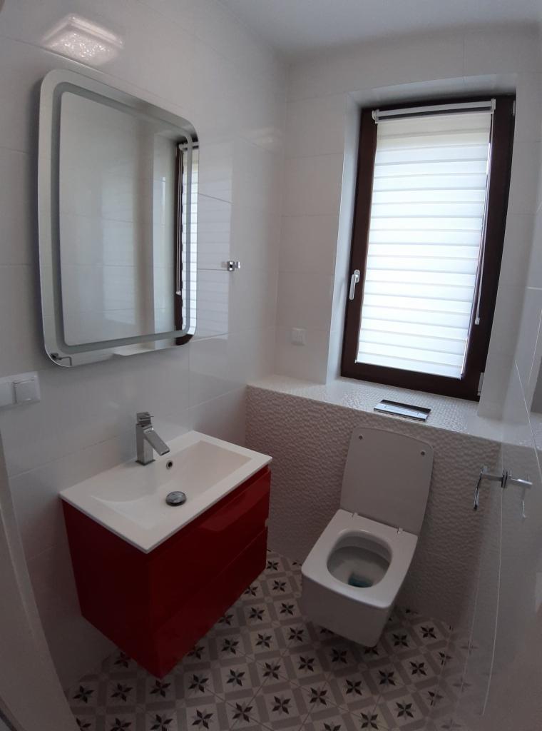 Nowoczesna łazienka z czerwona szafką pod umywalką, toaletą z deską wolnoopadającą i lustrem z podświetleniem LED, wykończona białymi płytkami i dekoracyjną mozaiką przy oknie z żaluzjami.