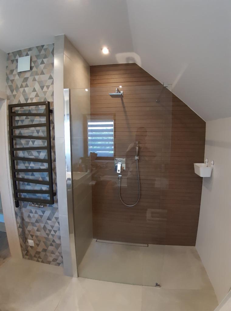 Nowoczesna kabina prysznicowa z deszczownicą i panelem prysznicowym na ścianie z drewnopodobnych paneli, obok grzejnik drabinkowy na ścianie z trójkątnych płytek w odcieniach szarości.