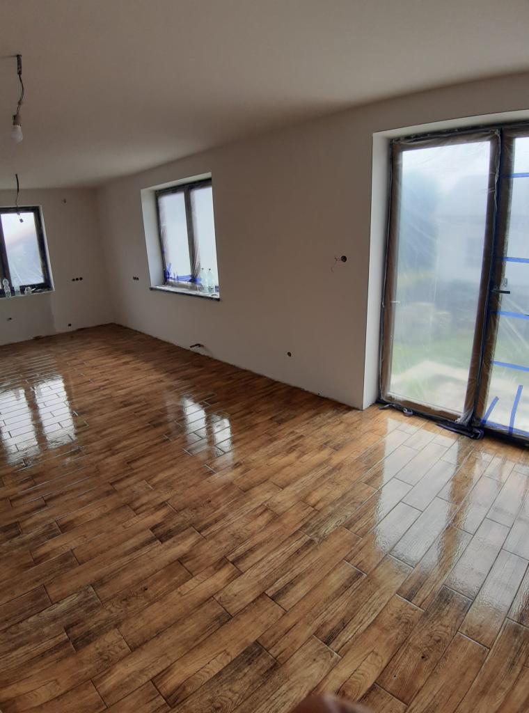 Nowo położona, błyszcząca podłoga z płytek imitujących drewno w jasnym pomieszczeniu, okna zabezpieczone folią malarską, surowa ściana z widocznymi punktami po instalacjach.